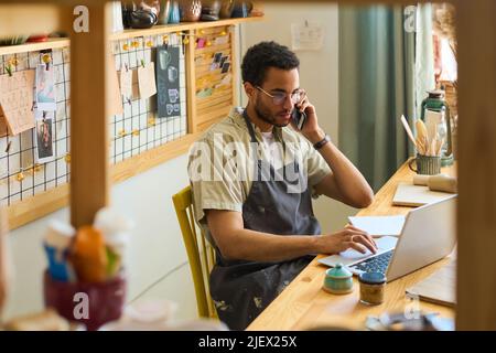 Young male clerk in apron calling clients and looking through online orders while sitting by workplace in front of laptop Stock Photo