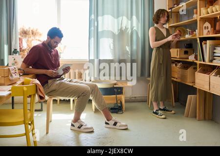 Guy with tablet working in the net while young woman standing by display and choosing handmade clay souvenir or mug Stock Photo