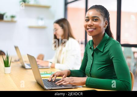 Portrait of smiling African American businesswoman with female colleague working on background, female office employees using laptops sitting in modern office space, diverse work team Stock Photo