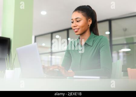 Portrait of concentrated african-american female employee using laptop computer, smiling black businesswoman responds emails, freelancer woman working on the distance sitting in coworking space Stock Photo