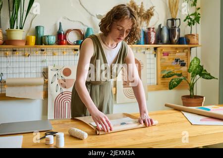 Young female clerk bending over table while wrapping self made abstract painting into paper while packing order of client in workshop Stock Photo