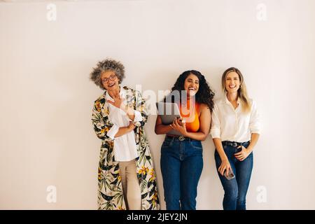 Three happy businesswomen laughing cheerfully while standing against a wall background. Group of multicultural businesswomen working together in an al Stock Photo