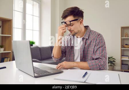 Sad tired overworked young man working on laptop rubs between eyes trying to remember something. Stock Photo