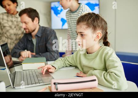Side view portrait of cute schoolgirl using laptop in modern school classroom with kids in background Stock Photo