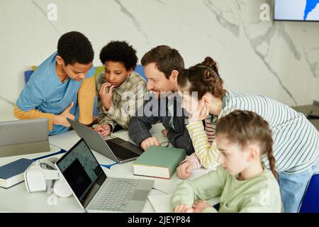 High angle portrait of smiling male teacher with diverse group of children using laptop together in school classroom Stock Photo