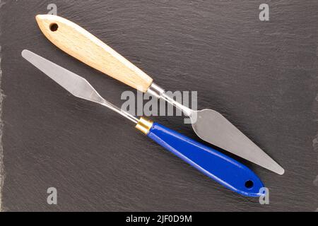 Two metal palette knives on a slate stone, close-up, top view. Stock Photo