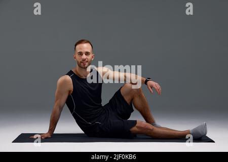 Smiling millennial guy doing abs exercises on sports mat over light studio background, panorama. Full length portrait of athletic man working out core Stock Photo