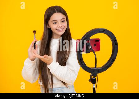 Beauty blog, presenting makeup cosmetics powder and brush. Teen girl speaking in front of camera for vlog isolated on yellow background. Teenager Stock Photo