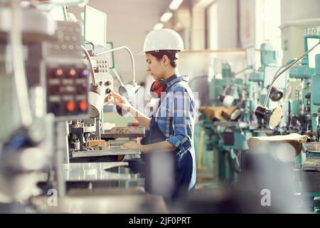 Busy young woman in overall standing in industrial workshop and operating manufacturing machine Stock Photo