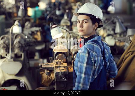 Portrait of serious young female production worker in hardhat standing at lathe and making watch movement Stock Photo