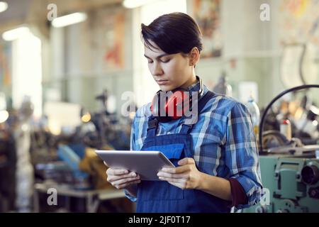Serious busy female factory engineer with short black hair standing in workshop and using tablet for lathe examination Stock Photo