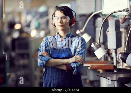 Portrait of serious professional female engineer in ear protectors and safety goggles standing at factory and crossing arms on chest Stock Photo