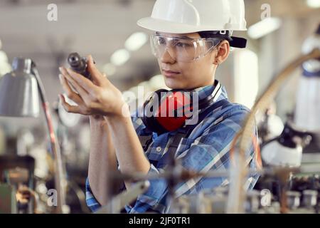 Serious busy young female repair engineer in hardhat and transparent goggles examining rotary detail at factory Stock Photo