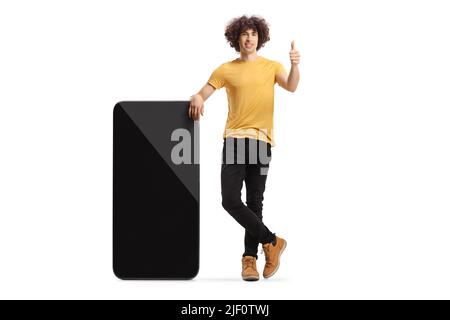 Young man with curly hair leaning on a smartphone and gesturing thumbs up isolated on white background Stock Photo