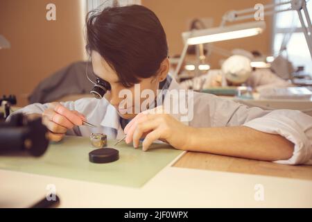 Concentrated young female watchmaker in lab coat sitting at table and using magnifying glass while assembling watch movements Stock Photo