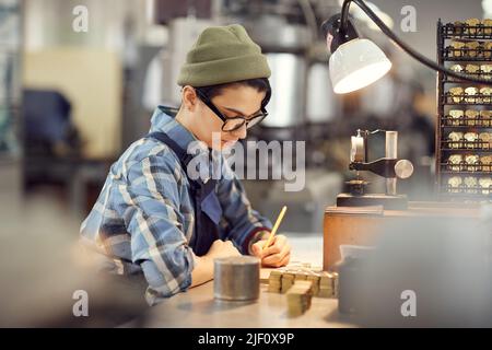 Serious concentrated hipster female watchmaker in beanie hat and glasses sitting at desk and making notes while working with watch movements Stock Photo