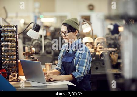 Serious skilled factory girl in eyeglasses sitting at table and making records on laptop while working at watch plant Stock Photo