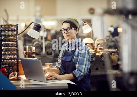 Portrait of content hipster female worker in eyeglasses sitting at desk and using laptop while composing manual for assembling wristwatch Stock Photo