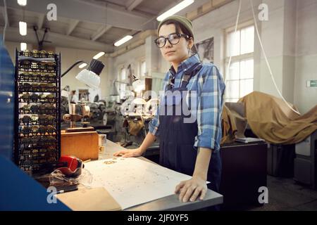 Portrait of serious female production worker in hipster hat standing at desk and assembling wristwatch movement according to scheme Stock Photo