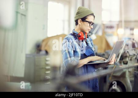 Serious busy young female engineer in hipster hat standing in factory shop and identifying industrial machine errors using laptop Stock Photo