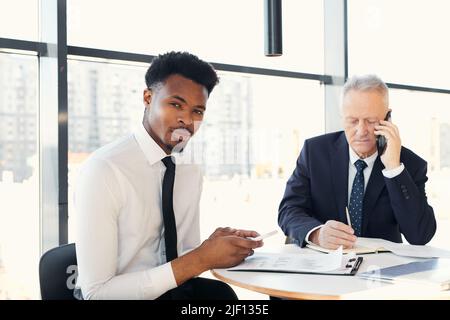Portrait of content confident young black businessman with beard using smartphone for notes while working on papers with mature colleague Stock Photo
