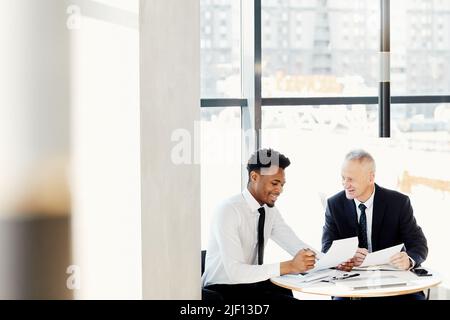 Smiling multi-ethnic business partners in formal outfits sitting at round table and viewing business papers together Stock Photo