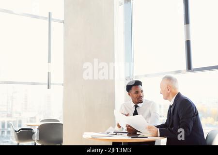 Two multi-ethnic businessmen in formal outfits sitting at table in modern office and doing paperwork together Stock Photo