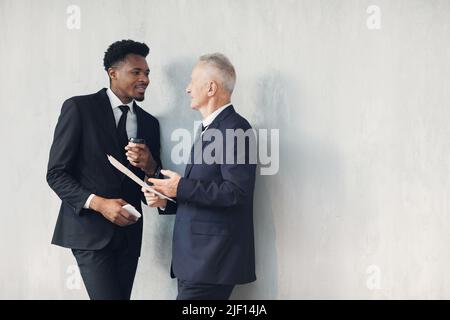 Two multi-ethnic business specialists in formal suits standing at wall and discussing financial issues while looking through papers Stock Photo