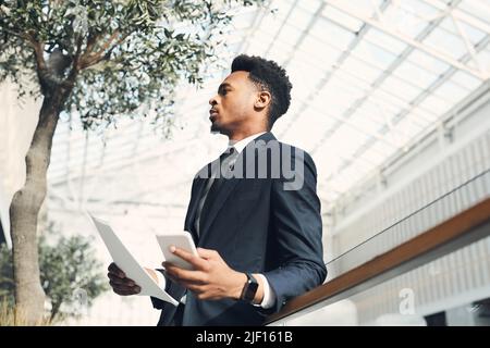 Serious young Afro-American manager in suit leaning on railing and using smartphone while thinking about sales analysis Stock Photo