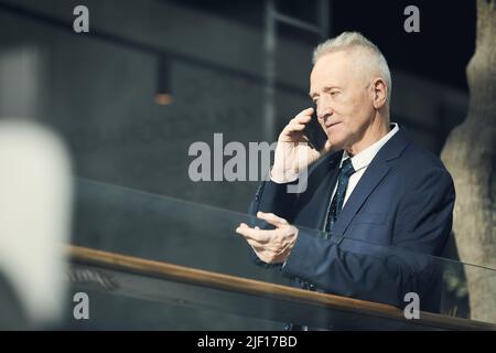 Confident senior businessman in formal jacket standing in modern lobby and gesturing hand while talking by cellphone Stock Photo