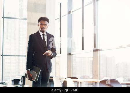 Brutal young black businessman in fashionable suit holding folders and using smartphone while standing in airport waiting area Stock Photo