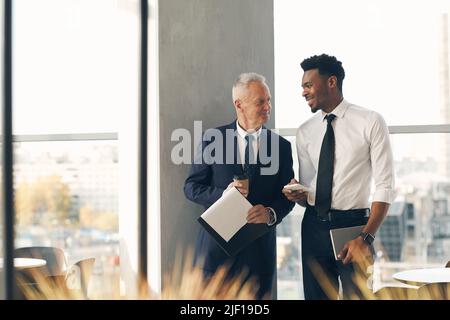 Positive multi-ethnic business colleagues in formalwear drinking coffee and chatting in lobby during break Stock Photo