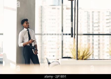 Dreamy handsome young African-American businessman in stylish formalwear standing in open space and using smartphone Stock Photo