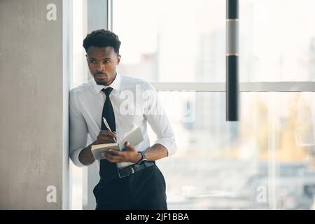 Serious pensive young Afro-American employee leaning on wall in hall and making notes in organizer while planning work day Stock Photo