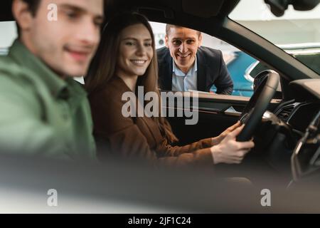 Young couple buying new car, sitting in auto salon, going on test drive, talking to salesman at automobile dealership Stock Photo