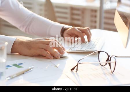 Close-up of unrecognizable male analyst in white shirt sitting at table in office and using computer while researching internet market Stock Photo