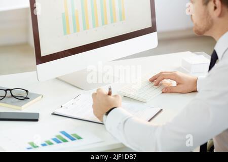 Over shoulder view of busy young market analyst using modern computer and making notes in report while analyzing graph Stock Photo