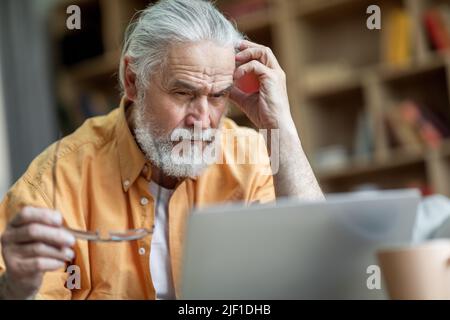 Pensive old man using notebook at home, touching head Stock Photo