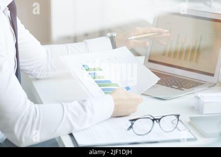 Close-up of unrecognizable market analyst sitting at desk and comparing graphs in laptop and papers while working in office, behind glass view Stock Photo