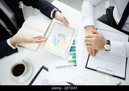 Above view of unrecognizable market analysts sitting at table in office and viewing pie chart on tablet while discussing sales changes Stock Photo