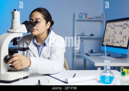 Serious young Indian female chemist in lab coat sitting at desk and using microscope in laboratory office Stock Photo