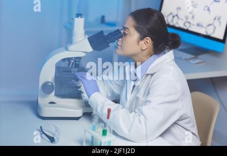 Concentrated young Indian female lab technician in white coat sitting at desk and working with microscope in modern laboratory Stock Photo