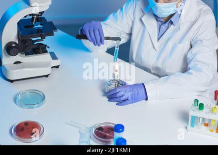 Close-up of unrecognizable lab technician in gloves and mask sitting at desk and using burner to catalyze chemical reaction Stock Photo