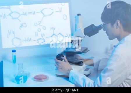 Serious focused modern Asian scientist in glasses sitting at desk in laboratory and using microscope while studying molecules Stock Photo