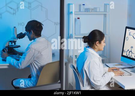 Busy young multi-ethnic chemists in lab coats sitting at tables in modern laboratory and working on research Stock Photo