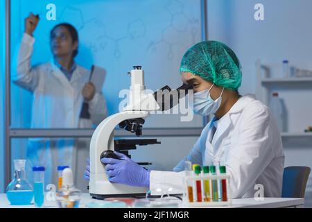 Concentrated young scientist in surgical cap and white cap sitting at desk and using microscope while working on chemical research in laboratory Stock Photo