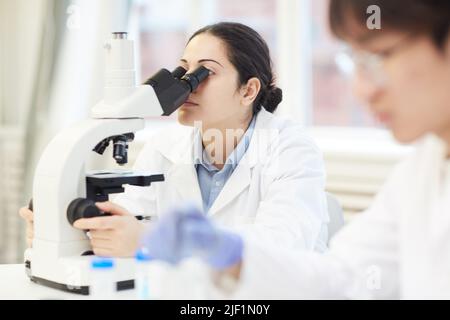 Focus on concentrated young Indian female biologist sitting at table and using microscope while examining sample Stock Photo