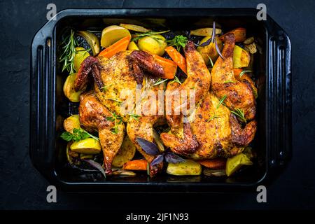 Baked whole chicken with potatoes, carrots and herbs in baking pan on black background Stock Photo