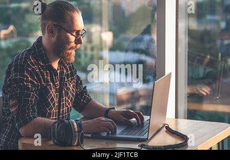A man with a beard works at a computer, sitting in the office by the window. Stock Photo