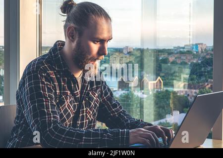 A man with a beard works at a computer, sitting in the office by the window. Stock Photo